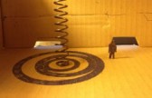 Detail of a cardboard model maquette for a sculpture. A spiral is drawn of the floor of the model, and a metal spring stands at the centre of the spiral.  A small cardboard human figure is next to the spiral.